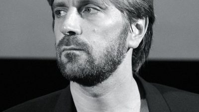 Ruben Östlund sns från axlarna ch uppåt. Han har halvlångt hår, skägg ch svart kavaj.
