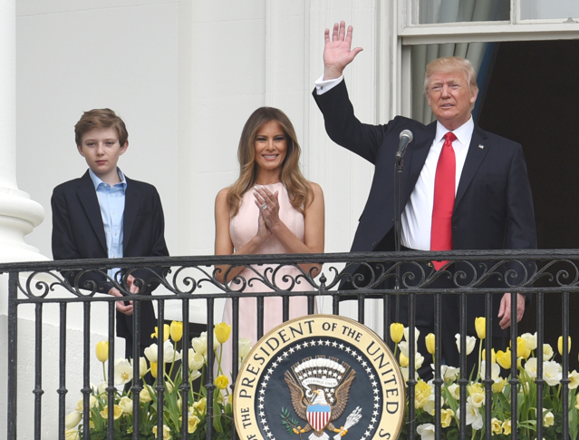 Donald Trum står på vita husets balkong och vingar med en hand. Bredvid honom står Melania som klappar händerna. Bredvid Trump och Melania står deras son.