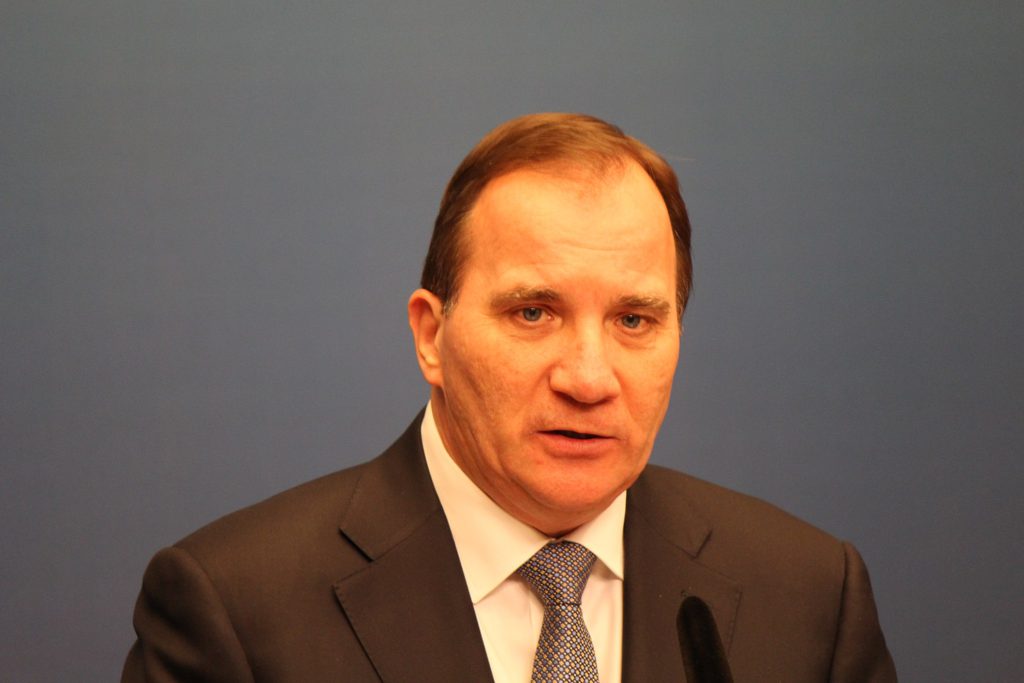 Stefan Löfven står och pratar med en mikrofon framför sig. Han har kavaj, skjorta och slips. Han har tunt brunt hår, små ögon och en stor näsa.