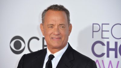 Tom Hanks står fram en vit vägg. Han har kort hår som är lite grått på sidorna. Han har kostym och slips på sig.