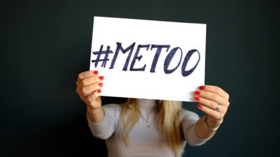 En tjej står mot en svart vägg. Vi ser inte hennes ansikte för framför sig sträcker hon fram en skylt där det står #metoo.