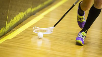 Fötter med sportskor springer över ett trägolv. Bredvid fötterna syns en bandyklubba som föser en bandyboll framför sig.