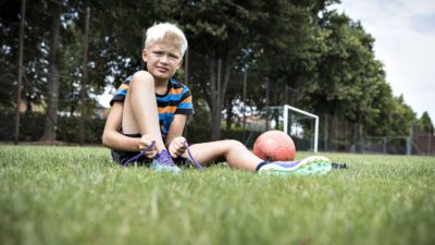 Julle sitter på en gräsmatta. Han knyter sina fotbollskor. Bredvid honom ligger en fotboll. I bakgrunden syns ett fotbollsmål.