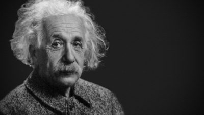 Albert Einstein sitter och tittar mot kameran. Han har stort rufsigt vitt hår, en stor mustasch och ganska många rynkor i pannan. Han ser snäll men lite trött ut.