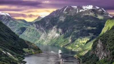 Bild på en fantastik utsikt över en fjord i Norge. Det är solnedgång bakom bergen och i vattnet ligger två stora båtar.